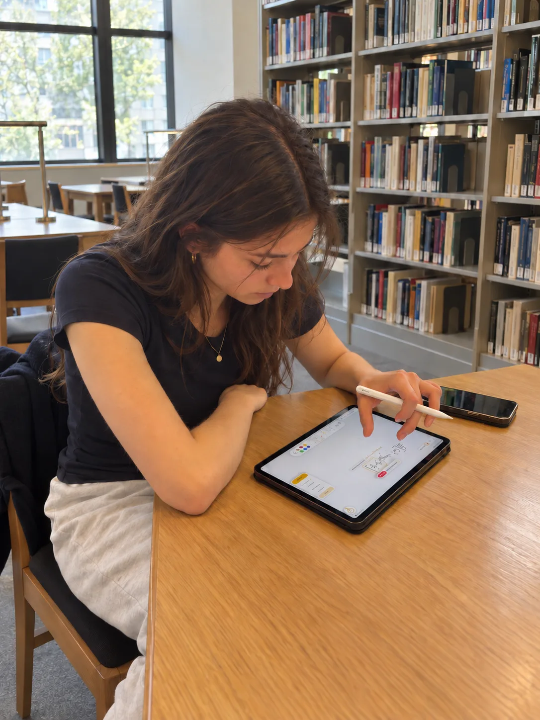 A student in a university library working on an iPad with Apple Pencil, Klera open on the screen